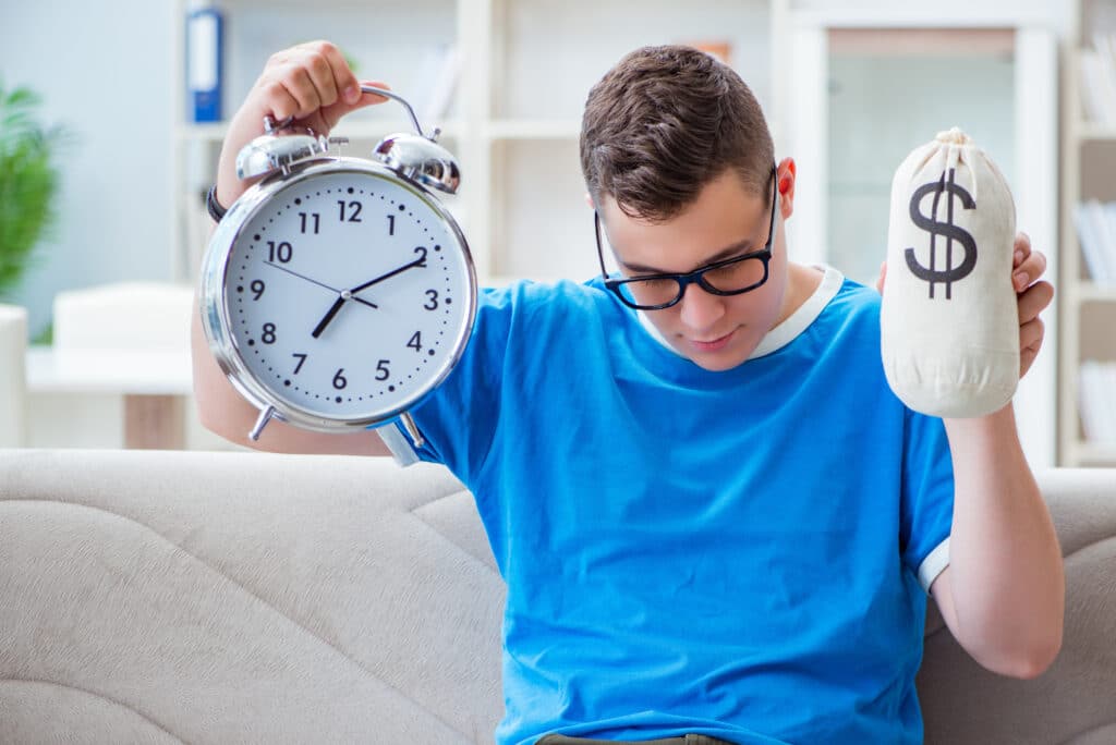 Young student preparing for exams studying at home on a sofa A young person looking busy and holding up a large clock with one hand and money in the other.