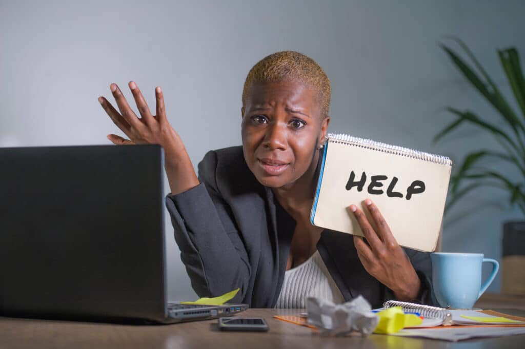 sad and depressed black afro American woman suffering stressed a Afircan-American woman at desk with computer and sign with "Help" written on it.